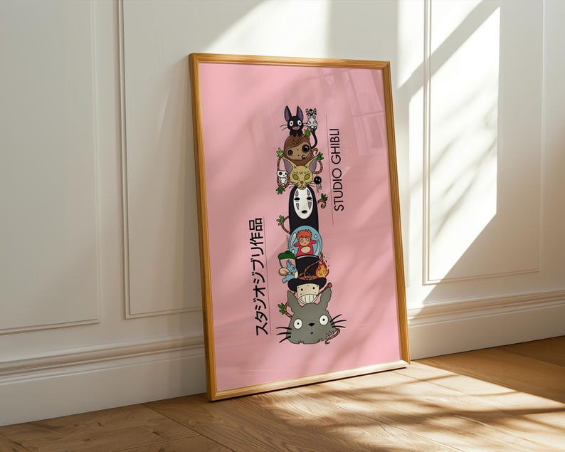 Puede incluir: Un p&oacute;ster rosa enmarcado con un marco dorado, que presenta una pila de personajes de Studio Ghibli. El p&oacute;ster tiene texto japon&eacute;s. Los personajes incluyen a Totoro y Sin Rostro. El p&oacute;ster est&aacute; apoyado contra una pared blanca y un suelo de madera.