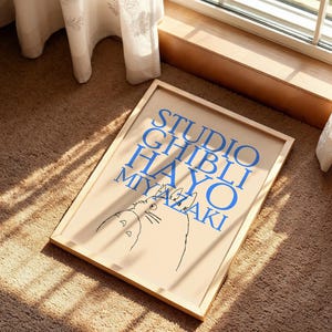 K&ouml;nnte beinhalten: Ein gerahmtes Poster mit dem blauen Text "STUDIO GHIBLI HAYAO MIYAZAKI" und einer Katzenzeichnung. Das Poster befindet sich in einem hellen Holzrahmen und liegt auf einem braunen Teppich in der N&auml;he eines Fensters.