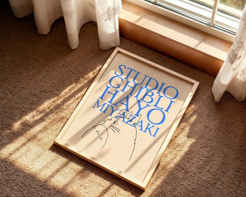 K&ouml;nnte beinhalten: Ein gerahmtes Poster mit dem blauen Text "STUDIO GHIBLI HAYAO MIYAZAKI" und einer Katzenzeichnung. Das Poster befindet sich in einem hellen Holzrahmen und liegt auf einem braunen Teppich in der N&auml;he eines Fensters.