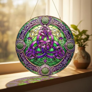 May include: A round stained glass window with a Celtic knot design. The window features a central purple knot, surrounded by green and purple accents, and a gold-colored metal frame. The window is hanging in front of a window.