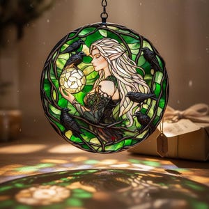 May include: Stained glass round panel featuring a woman with long white hair, surrounded by black crows and green foliage. The woman holds a glowing orb. The panel is framed by a dark metal ring, creating a mystical and artistic piece.