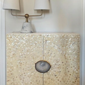 Könnte beinhalten: Ein weißer Schrank mit zwei Türen und einem dekorativen Achat-Knopf. Der Schrank ist aus Perlmutt gefertigt und hat ein einzigartiges, wirbelndes Muster. Der Schrank steht auf einer weißen Oberfläche und hat eine goldene Lampe mit zwei weißen Lampenschirmen darauf.