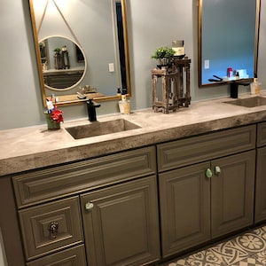 May include: A bathroom vanity with a concrete countertop and two sinks. The vanity has dark brown cabinets with green stone drawer pulls. The countertop has a decorative wooden stand with a plant and candles.