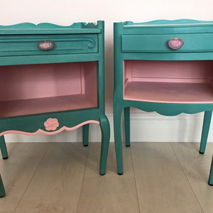 May include: Two teal blue bedside tables with pink interiors and decorative floral accents. Each table has a drawer with a floral knob and a lower shelf.