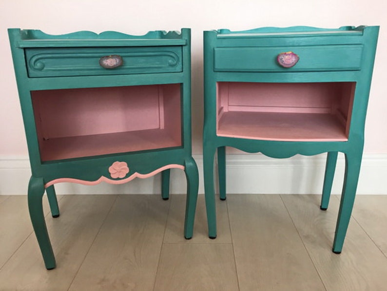May include: Two teal blue bedside tables with pink interiors and decorative floral accents. Each table has a drawer with a floral knob and a lower shelf.