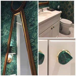May include: A bathroom with green and gold wallpaper, a white vanity with a white countertop, a white toilet, and a gold-framed mirror. The vanity has gold knobs with green malachite stone accents.