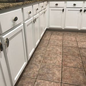 May include: White kitchen cabinets with agate drawer pulls. The cabinets are arranged in a row and have a light wood finish. The floor is tiled in a brown and beige pattern.