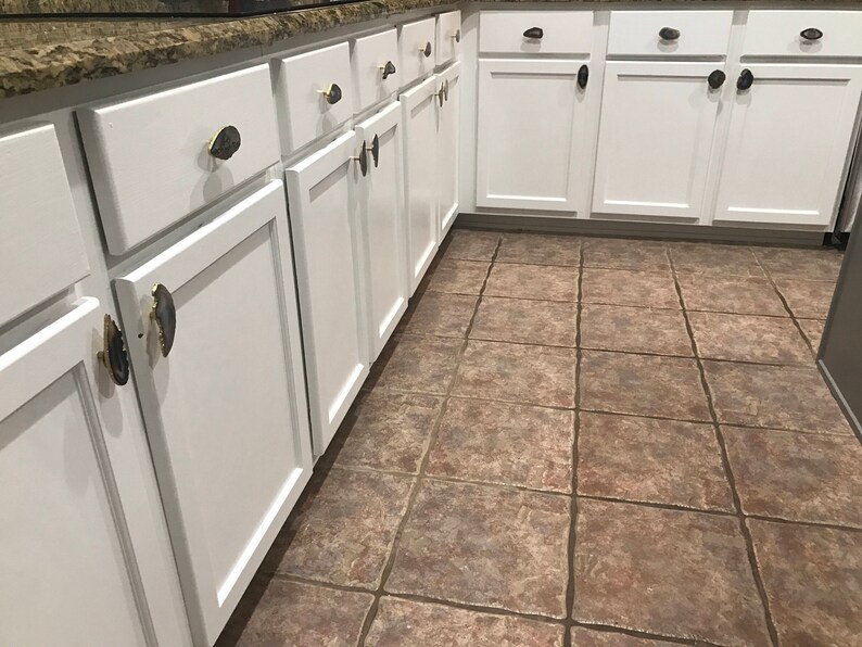 May include: White kitchen cabinets with agate drawer pulls. The cabinets are arranged in a row and have a light wood finish. The floor is tiled in a brown and beige pattern.