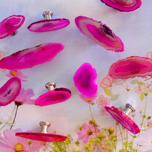 May include: Nine pink agate slices with silver knobs, arranged on a white surface with a floral pattern. The agate slices are various sizes and shapes, with a natural, organic look.