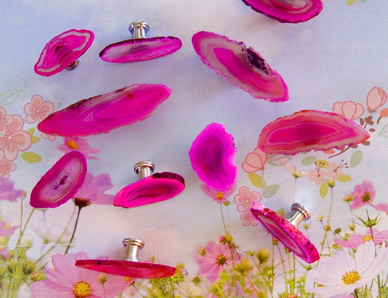 May include: Nine pink agate slices with silver knobs, arranged on a white surface with a floral pattern. The agate slices are various sizes and shapes, with a natural, organic look.
