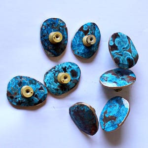 Könnte beinhalten: Eine Sammlung von acht dekorativen Schrankknöpfen. Jeder Knopf hat eine einzigartige, unregelmäßige Form mit einem lebendigen blau-braunen Muster, akzentuiert durch einen goldfarbenen Rand und eine Messingschraube. Diese Knöpfe sind ideal, um Möbeln einen Hauch von künstlerischem Flair zu verleihen.