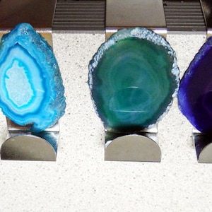 May include: Seven agate slices in different colors, including blue, green, and purple, are displayed on metal stands. The agate slices are polished and have a natural, organic look.