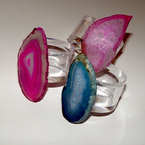 May include: Three agate napkin rings, one pink, one blue, and one clear, with a white background.