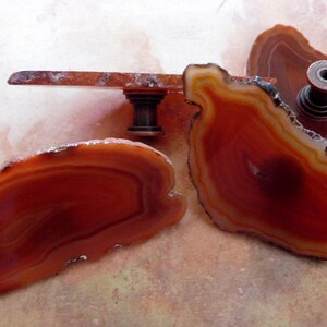 May include: Three agate slices in shades of brown and orange with a dark brown metal knob attached to one of the slices.