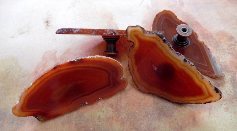 May include: Three agate slices in shades of brown and orange with a dark brown metal knob attached to one of the slices.