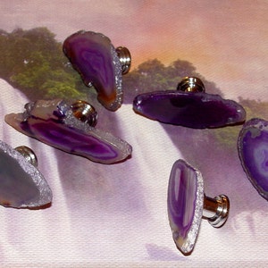 May include: Six purple agate drawer pulls with silver metal hardware. The agate is translucent and has a variety of purple and white patterns.