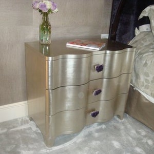 May include: A silver bedside table with three drawers and decorative purple crystal drawer pulls. The table has a wavy, curved design.