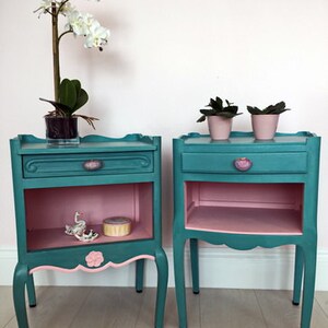 May include: Two teal blue bedside tables with pink accents. Each table has a drawer and a shelf. The tables are on a light wood floor.