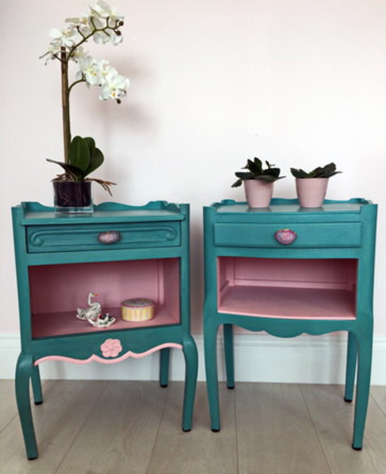 May include: Two teal blue bedside tables with pink accents. Each table has a drawer and a shelf. The tables are on a light wood floor.
