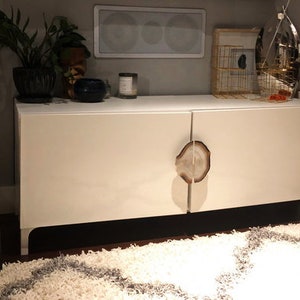 Könnte beinhalten: Ein weißer, moderner Schrank mit zwei Türen und einzigartigen, natürlich aussehenden Griffen. Die Griffe scheinen Scheiben eines Baumstamms zu sein. Der Schrank steht vor einer neutralen Wand, mit einem Teppich im Vordergrund und dekorativen Gegenständen.