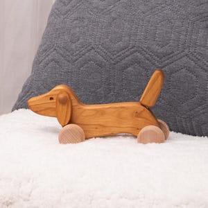 Può includere: Un giocattolo in legno a forma di bassotto con finitura marrone chiaro. Il giocattolo ha un corpo lungo, orecchie flosce e una coda sollevata. Poggia su quattro ruote di legno. Il giocattolo è posto su una coperta bianca e soffice, con un cuscino grigio trapuntato sullo sfondo.