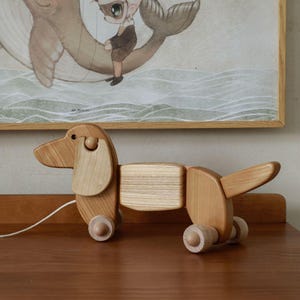Könnte beinhalten: Ein Dackel-Spielzeug aus Holz in hellem Braun. Das Spielzeug hat einen segmentierten Körper, schlaffe Ohren und kleine Holzräder. Ein weißes Kabel erstreckt sich von dem Spielzeug. Das Spielzeug steht auf einer Holzoberfläche, mit einer gerahmten Illustration im Hintergrund.