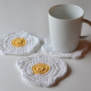 Puede incluir: Tres posavasos de ganchillo blanco con forma de huevo frito con centros amarillos. Una taza de cerámica blanca se encuentra sobre uno de los posavasos.