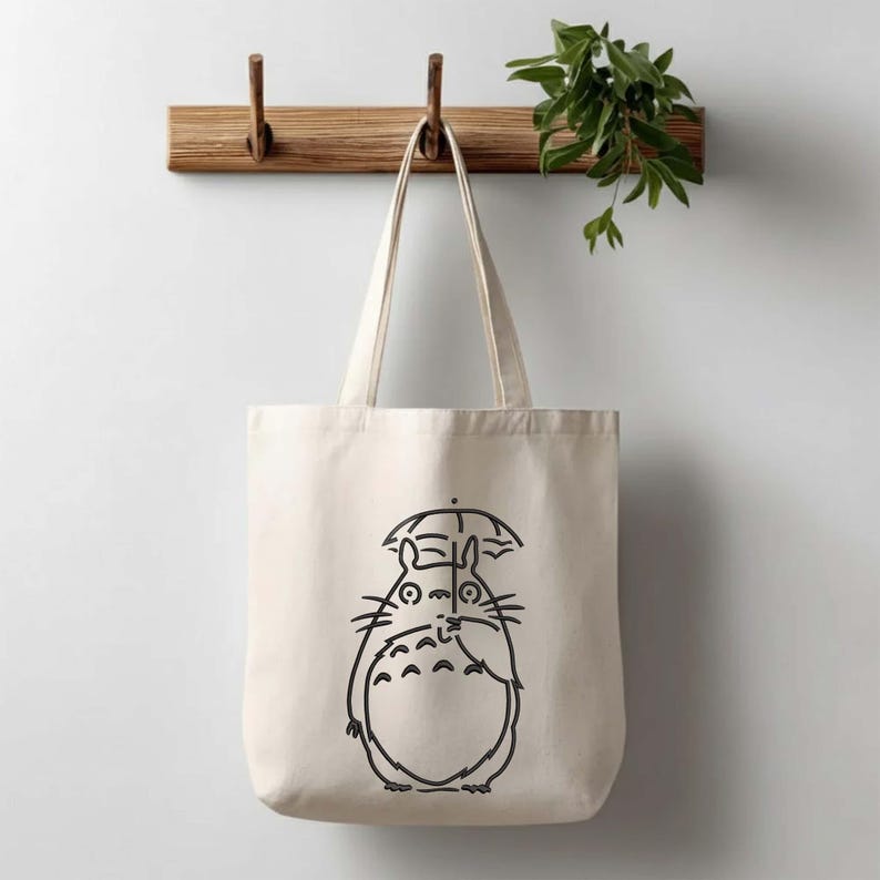 May include: A cream-colored tote bag with a black line drawing of a Totoro character holding an umbrella. The bag has two handles and is hanging on a wooden rack. A green plant is visible in the background.