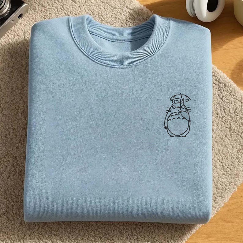 May include: A light blue sweatshirt with a crew neck and a small black graphic of a cartoon character holding an umbrella. The sweatshirt is folded neatly on a beige, textured surface.