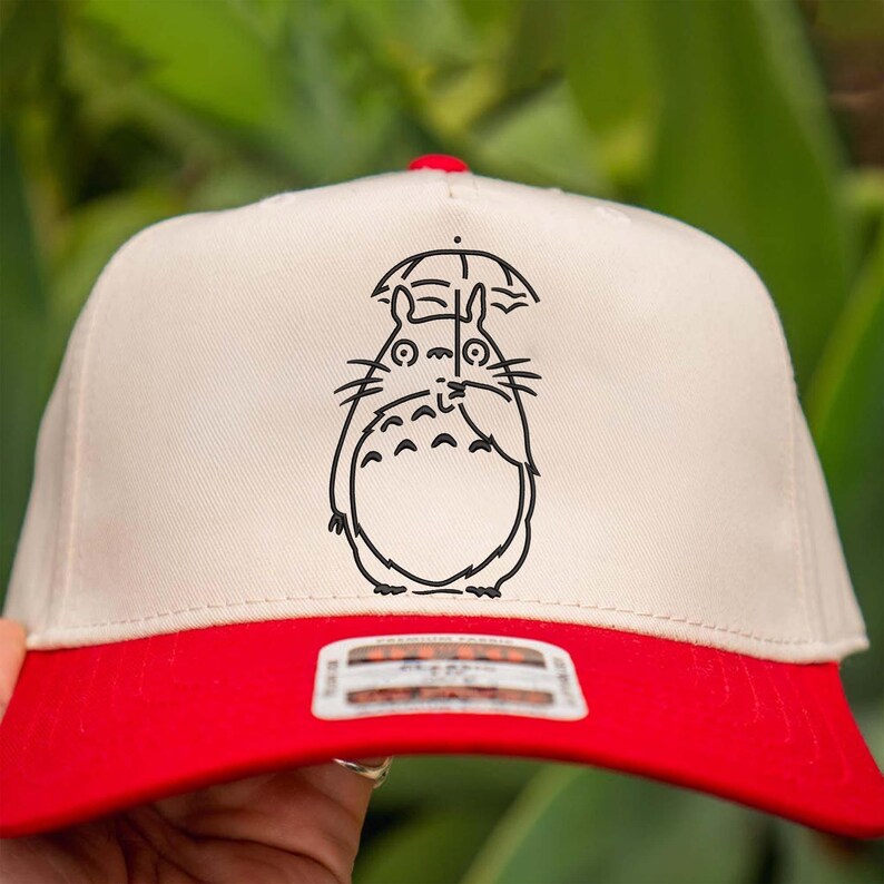 May include: A baseball cap with a cream-colored front panel and a red brim. The cap features a black graphic of a cartoon character holding an umbrella. The cap is a classic style, suitable for casual wear.