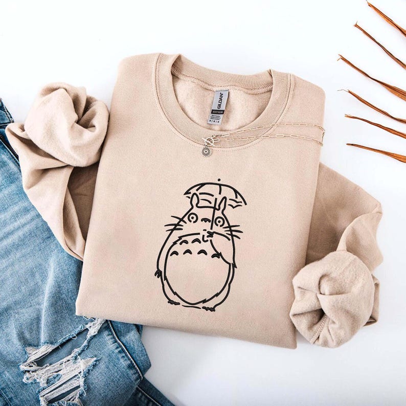 May include: Beige crewneck sweatshirt featuring a black graphic of a cartoon character with an umbrella. A silver necklace with a small pendant is draped over the sweatshirt. The sweatshirt is folded, with a pair of blue jeans visible in the background.