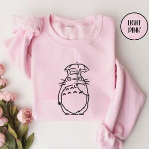 May include: Light pink sweatshirt featuring a black line drawing of a cartoon character holding an umbrella. The sweatshirt has long sleeves and a crew neck. The words "LIGHT PINK" are in a circle in the upper right corner.