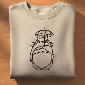May include: A beige sweatshirt featuring a black line drawing of a cartoon character with an umbrella. The character has a large, round body, small arms, and a smiling face. The sweatshirt is folded neatly.
