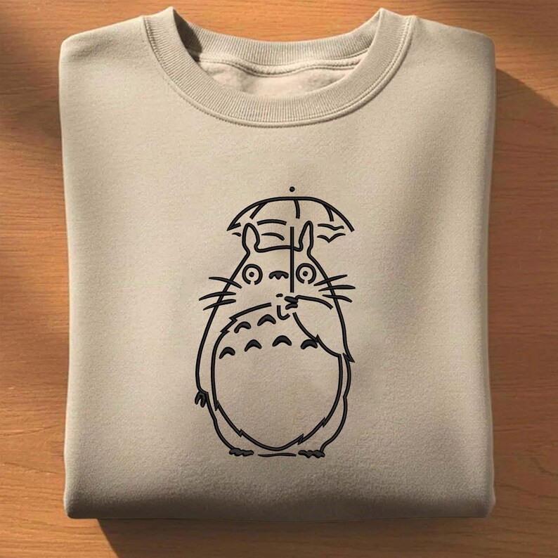 May include: A beige sweatshirt featuring a black line drawing of a cartoon character with an umbrella. The character has a large, round body, small arms, and a smiling face. The sweatshirt is folded neatly.