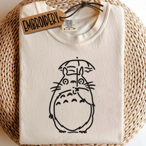 May include: Cream-colored t-shirt featuring a black outline of a cartoon character holding an umbrella. The shirt has a tag that says "EMBROIDERY". The shirt is folded and placed on a woven surface. The shirt is a Comfort Colors brand, size S-P-CH.