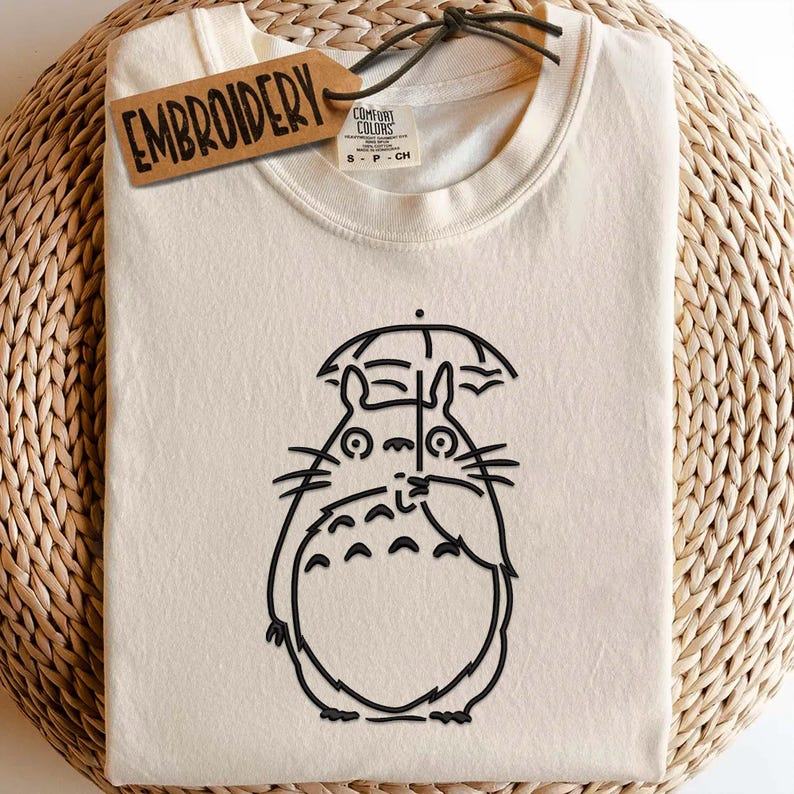 May include: Cream-colored t-shirt featuring a black outline of a cartoon character holding an umbrella. The shirt has a tag that says "EMBROIDERY". The shirt is folded and placed on a woven surface. The shirt is a Comfort Colors brand, size S-P-CH.