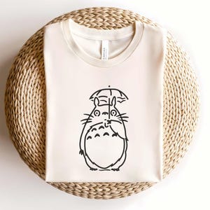 May include: Cream-colored t-shirt featuring a black line drawing of a cartoon character holding an umbrella. The character has large eyes, whiskers, and a round body. The shirt is made of a soft material and is displayed on a woven surface.