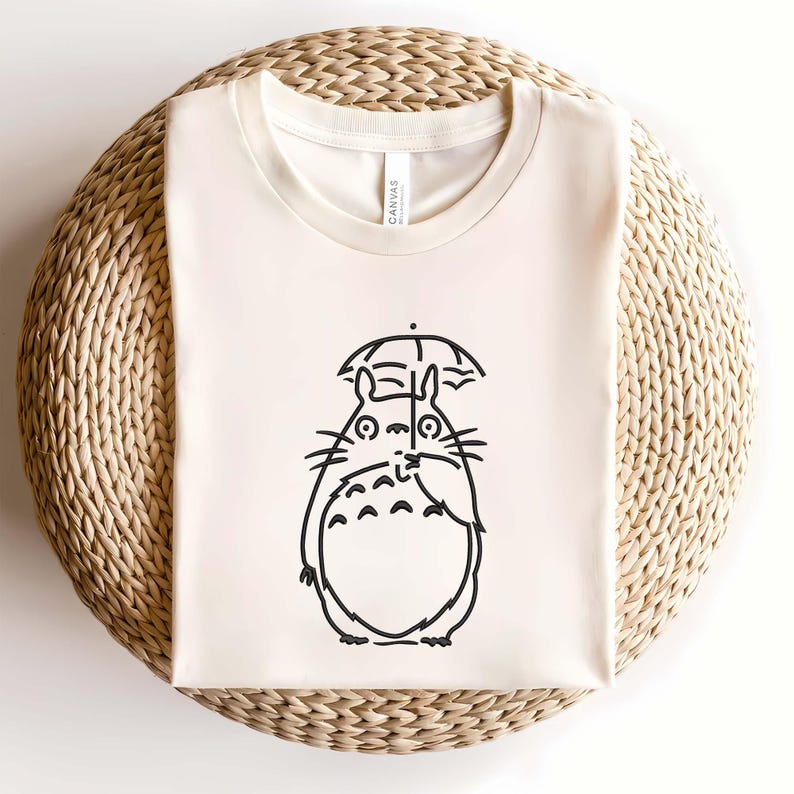 May include: Cream-colored t-shirt featuring a black line drawing of a cartoon character holding an umbrella. The character has large eyes, whiskers, and a round body. The shirt is made of a soft material and is displayed on a woven surface.