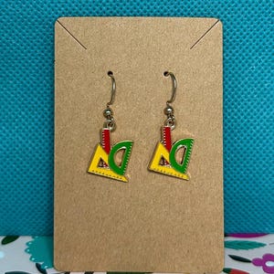 Puede incluir: Un par de pendientes colgantes con un diseño de regla y transportador. Los pendientes tienen un triángulo amarillo, un transportador verde y una línea vertical roja. Los pendientes se muestran en una tarjeta marrón.