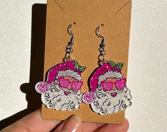 May include: A pair of Santa Claus earrings with pink sunglasses and hats. The earrings have silver hooks and are displayed on a brown card. The Santa faces have white beards and glitter accents, perfect for the holiday season.