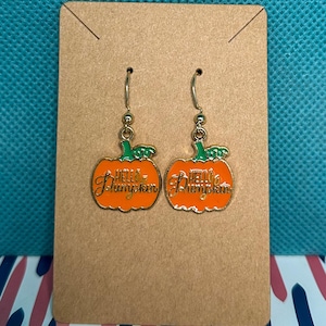 May include: A pair of orange pumpkin earrings with the text "Hello Pumpkin" in gold lettering. The earrings have green stems and gold-colored hooks, displayed on a brown card.