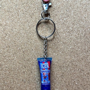 May include: A keychain featuring a miniature blue ICEE Squeeze Candy tube with the text "ICEE Squeeze Candy" and a graphic of a person. The keychain has a silver clasp, a chain, and a pink glittery bead.