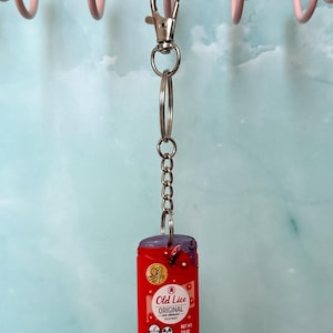 May include: A red and purple keychain featuring a miniature deodorant container with the text "Old Lice Original". The keychain has a silver clasp and chain, and the deodorant container has a graphic of a bug.