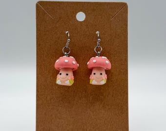 May include: A pair of dangle earrings featuring small, pink mushroom charms with white spots and smiling faces. The mushrooms have light pink stems with small yellow floral accents at the base. The charms hang from silver-toned metal earring hooks.