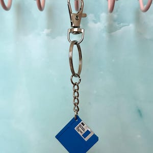 Puede incluir: Un llavero de disquete azul con un mosquetón y una cadena plateados. El disquete tiene una etiqueta blanca con texto negro y el logotipo de 3M. El llavero cuelga de un gancho rosa.