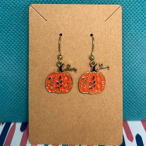 Puede incluir: Un par de pendientes de calabaza naranja con detalles dorados y un diseño floral. Los pendientes se exhiben en una tarjeta marrón sobre un fondo turquesa, perfectos para el otoño.
