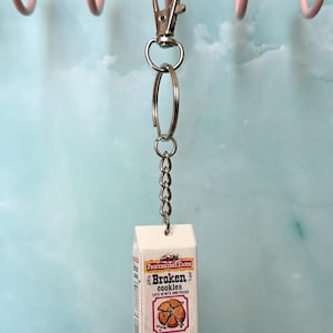May include: A miniature white carton keychain featuring the text Poopedridge Farm Broken cookies lots of bits and pieces and a graphic of a cookie. The carton includes the text net wt 5 1/2 crumbling oz and is attached to a silver metal ring.