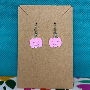 Puede incluir: Pendientes de calabaza rosa con ganchos plateados. Los pendientes se muestran en una tarjeta marrón sobre un fondo turquesa. Las calabazas tienen una cara sonriente y probablemente sean para Halloween.