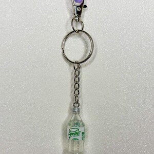 May include: A novelty keychain featuring a miniature Sprite bottle. The clear plastic bottle has a green and white label with the Sprite logo. The keychain includes a silver-colored chain, a key ring, and a purple clasp.