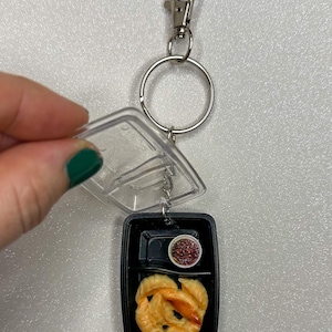 May include: Miniature food keychain featuring a black bento box with dumplings and a small dish of sauce. The keychain includes a silver ring, a clasp, and a purple bead.